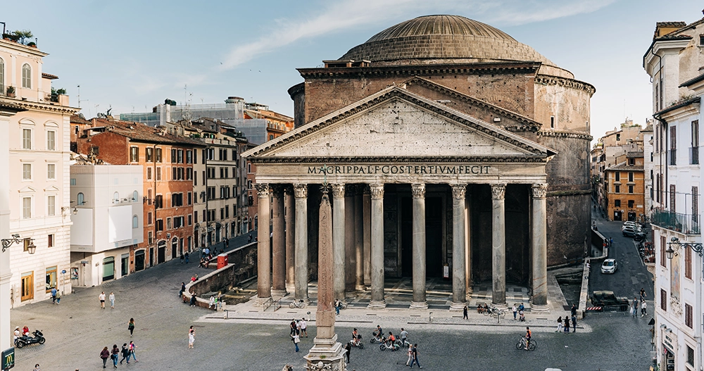 Roma'nın en iyi korunmuş antik yapılarından Pantheon, görkemli sütunlu girişi ve devasa kubbesiyle Piazza della Rotonda'da çevresindeki tarihi İtalyan binalarıyla birlikte, bir Roma seyahat rehberinin vazgeçilmez durağı olarak güneşli bir günde ziyaretçilerini ağırlıyor.
