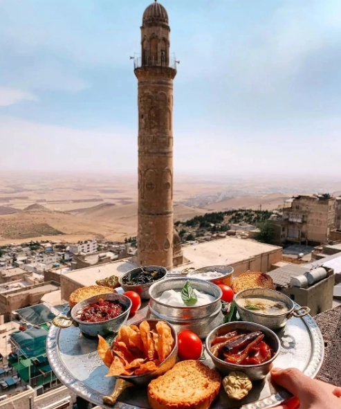 Gümüş bir tepside sunulan zeytin, yoğurtlu soslar, közlenmiş biber ve kızarmış patates gibi lezzetli atıştırmalıkların ön planda olduğu, arka planda ise tarihi Mardin'in eşsiz taş mimarisi, yüksek minaresi ve Mezopotamya Ovası'nın nefes kesen genel görünümüyle seyirlik bir manzara.