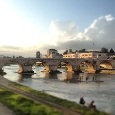 Gün batımı ya da doğumu ışıkları altında Vardar Nehri üzerinde uzanan, Üsküp'ün Tarihten Kalan İzleriyle öne çıkan simgesi Taşköprü, kemerli yapısıyla ve arkasındaki belirgin binalarla şehrin tarihi dokusunu yansıtırken, ön plandaki bulanık yeşil nehir kıyısı huzurlu bir atmosfer sunuyor.
