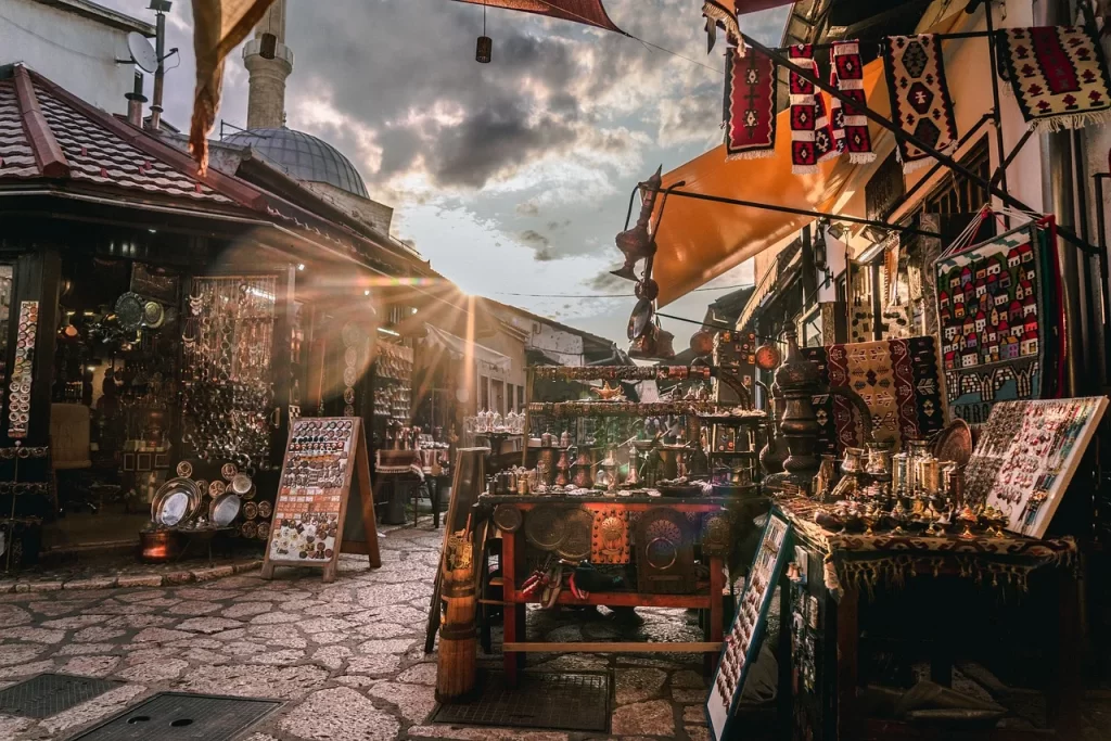 Üsküp'ün Tarihten Kalan İzleriyle dolu Eski Çarşı'sında, Arnavut kaldırımlı dar sokakta gün batımının parlak ışıkları altında geleneksel bakır eşyalar, el yapımı takılar ve renkli dokuma halılar satan dükkanlar sıralanırken, arka planda bir caminin minaresi ve kubbesi yükseliyor.