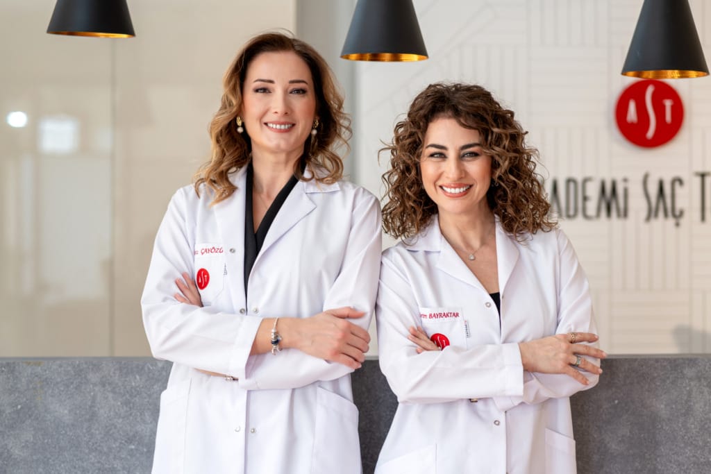 Akademi Saç Terapi merkezinin kurucuları Burcu Çayözü ve Evrim Bayraktar, soğuk havalarda saç bakımı üzerine uzman görüşlerini paylaşırken, beyaz önlükleriyle kameraya gülümseyerek poz veriyor.