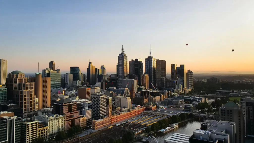 Gün doğumu veya batımının altın rengi ışıklarıyla aydınlanan Melbourne şehir manzarasında, modern gökdelenler ve geleneksel binalar kentsel dokuyu oluştururken, gökyüzünde süzülen iki sıcak hava balonu ve nehir kenarında uzanan hareketli tren istasyonu dikkat çekiyor.