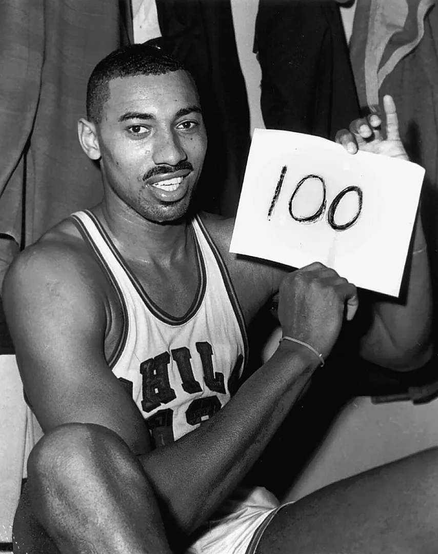 Basketbolun efsanevi oyuncusu Wilt Chamberlain, yüzünde gururlu bir gülümsemeyle kameraya bakarken, tarihi 100 sayılık rekorunu simgeleyen büyük 100 rakamının yazılı olduğu bir kağıdı sol elinde tutuyor.
