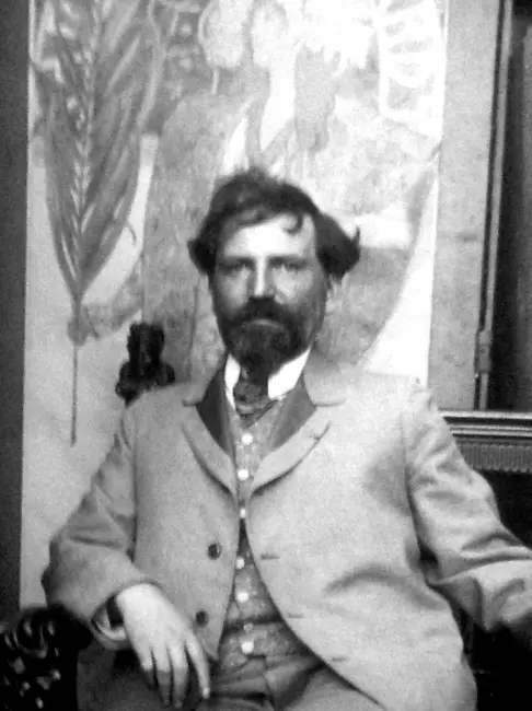 Alphonse Mucha’nın gençliğinde stüdyosunda çekilmiş siyah-beyaz portresi, arka planda sanatına ilham veren desenler ve figürler.