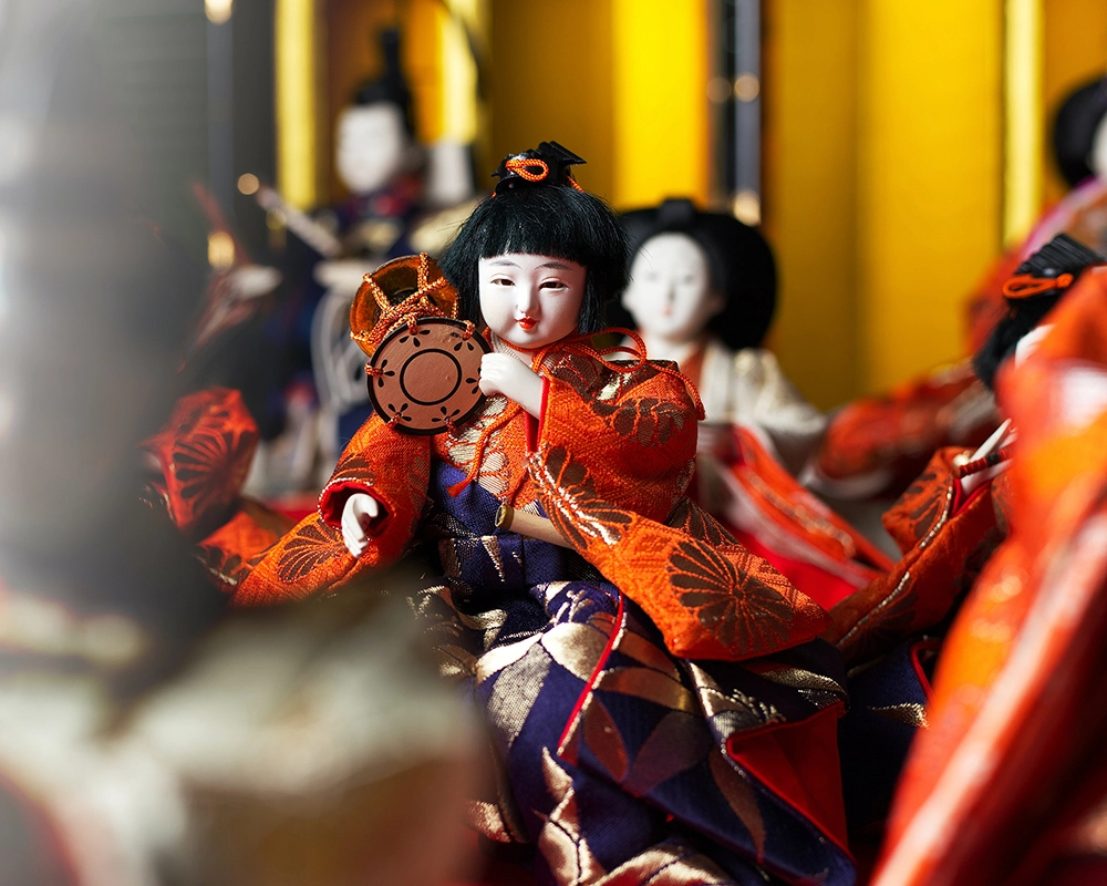 Geleneksel Japon kıyafetleri giymiş bir bebek figürü, elinde küçük bir enstrüman tutuyor; arka planda benzer bebek figürleriyle birlikte canlı renklerle öne çıkıyor.