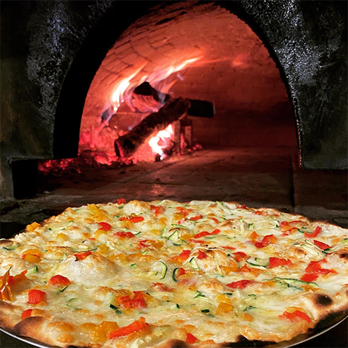 Ön planda taze mozzarella, kırmızı ve sarı biber dilimleri ile yeşil kabak parçacıklarıyla süslenmiş, kenarları çıtırlaşmış bir pizza metal bir tepside dururken, arka planda geleneksel taş fırının odun ateşi canlı alevlerle parlayarak Roma'nın eşsiz yemek kültürünü sergiliyor.