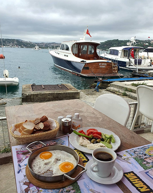 Bebek'in denize sıfır eşsiz Boğaz manzaralı bir mekanında, iki göz yumurta, taze sebzeler, peynir, ekmek ve kahve içeren zengin bir kahvaltı masası, arka planda demirli tekneler ve tepelerle huzurlu bir sabah keyfini yansıtıyor.