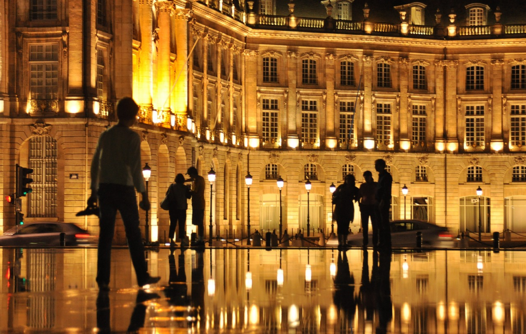 Gece ışıklarıyla aydınlatılmış Bordeaux'daki görkemli Place de la Bourse binasının ve etraftaki insanların siluetlerinin, ıslak zeminin ya da ünlü su aynasının üzerinde büyüleyici bir şekilde yansıdığı atmosferik bir görüntü.