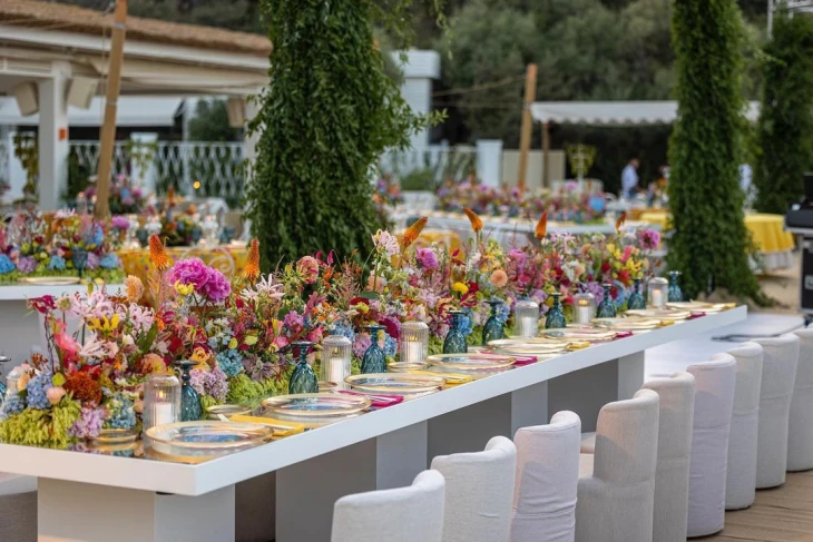 Açık hava yaz düğününde, canlı renkte çiçeklerle bezeli modern masa dekorasyonu; sarı, pembe, mor tonlarının hakim olduğu neşeli ve enerjik bir stil.