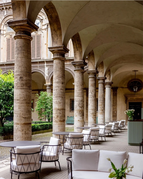 Roma seyahatinizde Via del Corso üzerinde konumlanan, tarihi Palazzo Doria Pamphilj'nin etkileyici iç avlusunda, yüksek kemerli taş sütunların gölgesinde sıralanan beyaz minderli modern oturma grupları ve masalarıyla hem kahve keyfi hem de sakin bir mola için ideal, huzurlu bir kafe ortamı.
