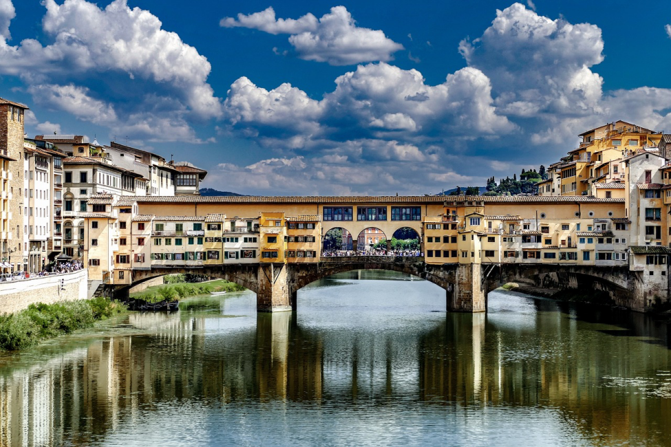 Floransa'daki tarihi Ponte Vecchio köprüsü, üzerinde sıralanmış rengarenk binalarıyla Arno Nehri'nin sakin suları üzerine uzanıyor; gökyüzündeki parlak mavi ve beyaz bulutlar, köprünün ve nehrin yansımalarıyla bütünleşerek büyüleyici bir manzara sunuyor.