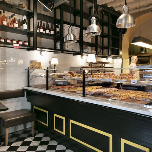 Roma seyahat rehberimizde gastronomi deneyimlerinin önemini vurgulayan, mermer tezgahta iştah açıcı kare kesim pizzaların ve çeşitli hamur işlerinin sergilendiği, raflarında İtalyan şarapları ve sosları bulunan geleneksel bir Roma fırınının çekici iç görünümü.