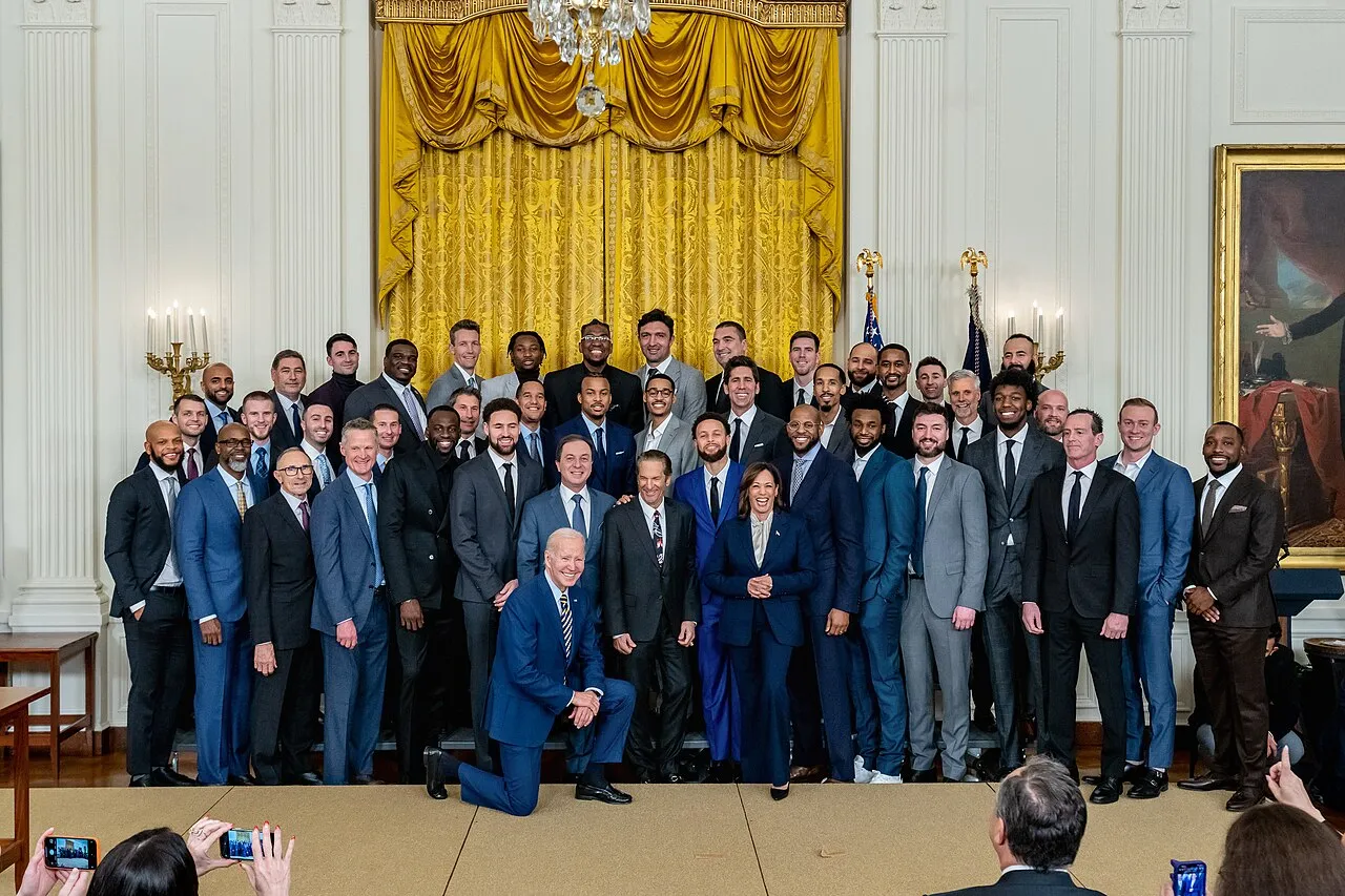 Golden State Warriors takımının oyuncuları, antrenörleri ve yöneticileri, NBA şampiyonluğu kutlamaları kapsamında Beyaz Saray'da Başkan Joe Biden'ın diz çöktüğü, Başkan Yardımcısı Kamala Harris ve yıldız oyuncu Stephen Curry'nin de yer aldığı, altın perdeler ve klasik sütunlarla süslü görkemli bir salonda çekilmiş bir grup fotoğrafında poz veriyor.