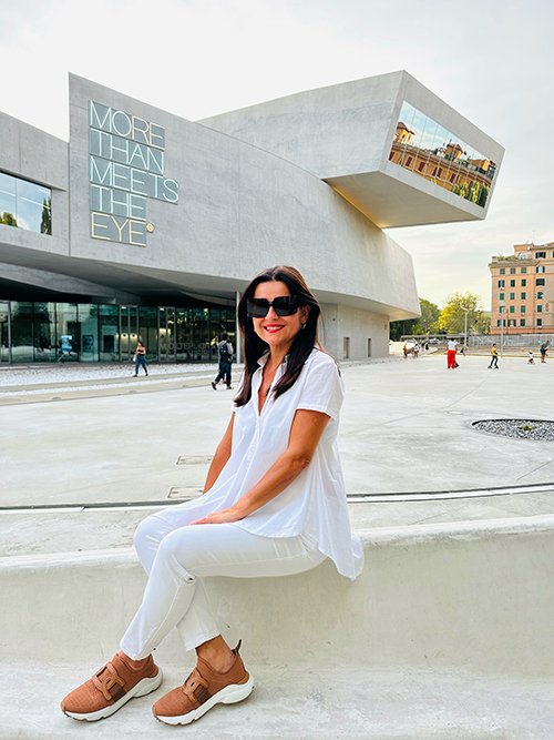 Güliz Özbek Collini, Roma'da modern mimarisiyle öne çıkan ve postun Çağdaş Sanat bölümünde önerilen Zaha Hadid imzalı Maxxi Müzesi'nin önündeki beton bir sette, beyaz kıyafetleri ve güneş gözlüğüyle gülümseyerek oturuyor, bu da Roma seyahat rehberi için keyifli ve güncel bir bakış açısı sunuyor.