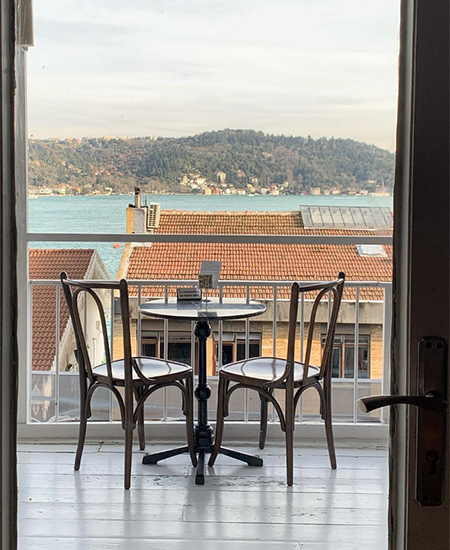 Bebek'teki en iyi mekanlardan birinin balkonundan, kırmızı kiremitli çatıların ve yemyeşil tepelerin arkasında uzanan masmavi Boğaz'ın büyüleyici manzarasına karşı yerleştirilmiş iki ahşap sandalyeli küçük bir masanın davetkar görüntüsü.