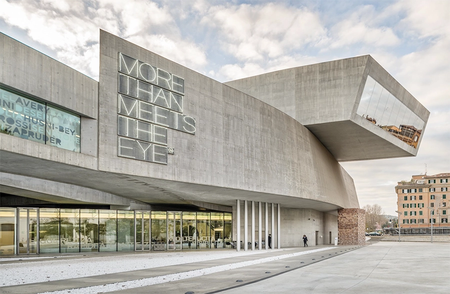 Roma seyahat rehberimizde yer alan, Zaha Hadid imzalı, dış cephesinde MORE THAN MEETS THE EYE yazısı ve açılı beton formlarıyla dikkat çeken modern Maxxi Çağdaş Sanat Müzesi, şehrin kültürel keşif duraklarından biri olarak gökyüzü yansımalarıyla parlıyor.