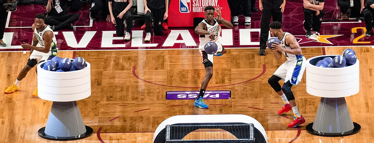 NBA All-Star etkinliğinde, Cleveland yazılı parkede üç basketbolcu yeteneklerini sergiliyor; solda sarı ayakkabılı oyuncu top dolu bir haznenin yanında dururken, ortadaki mavi ayakkabılı oyuncu topu ayağıyla dribble ediyor ve sağdaki kırmızı ayakkabılı oyuncu da topu tutuyor, bu anlar NBA All-Star Cumartesi Gecesi yarışmalarından birini yansıtıyor.