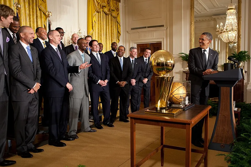 Beyaz Saray'da düzenlenen bir törende, eski Başkan Barack Obama, bir grup NBA şampiyonu oyuncu ve teknik ekiple birlikte güler yüzle poz veriyor, önlerindeki şık ahşap masada ise parıldayan devasa NBA şampiyonluk kupası ve imzalı bir basketbol topu sergileniyor.