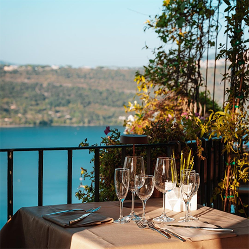 Ristorante Pagnanelli'nin balkonunda, Castel Gandolfo Gölü'nün nefes kesici manzarasına nazır, kahverengi bir masa örtüsü, çatal bıçak takımları ve çeşitli şarap kadehleriyle yemek için özenle hazırlanmış masa, Roma seyahati rehberinizdeki keyifli alternatifleri vurguluyor.