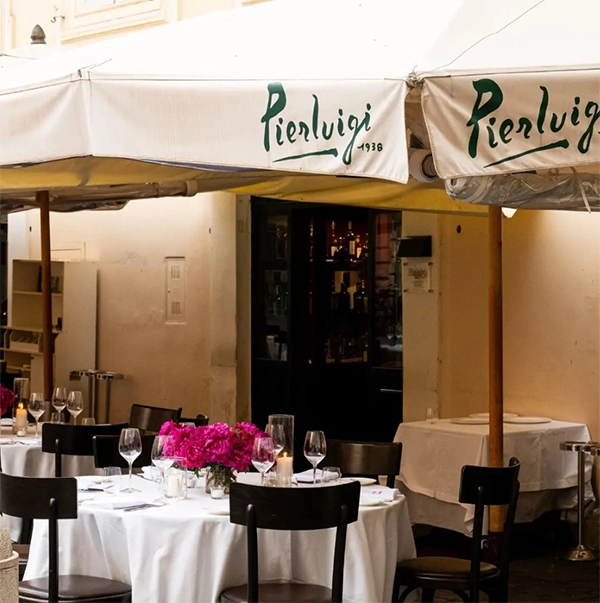 Pierluigi restoranının beyaz masa örtülü, pembe çiçeklerle ve şarap kadehleriyle donatılmış şık açık hava masaları, Roma seyahat rehberi kapsamında şehrin kaliteli gastronomi deneyimlerini sunan davetkar bir ambiyans sergiliyor.