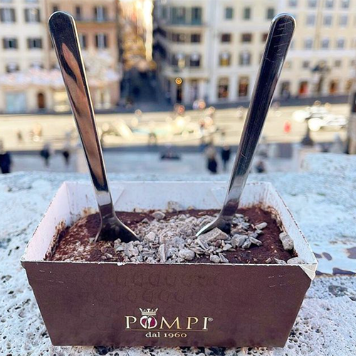 Pompi markalı kahverengi ambalajında, üzerinde kakao tozu ve küçük çikolata parçacıkları bulunan, içine iki metal kaşık batırılmış iştah açıcı bir tiramisu tatlısı, arka planda Roma'nın tarihi yapılarını anımsatan bulanık şehir manzarasıyla bu seyahat rehberinde bahsettiğimiz gastronomi deneyimini vurguluyor.