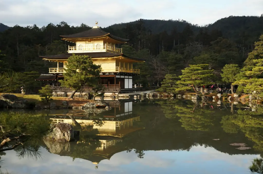 Kyoto, Japonya'da bulunan, yemyeşil bahçe ağaçları ve kayalarla çevrili sakin bir gölette parıldayan üç katlı altın rengi Kinkaku-ji tapınağının kusursuz yansıması, arka plandaki koyu yeşil ormanlık tepelerle huzurlu bir Japon manzarasını sergiliyor.