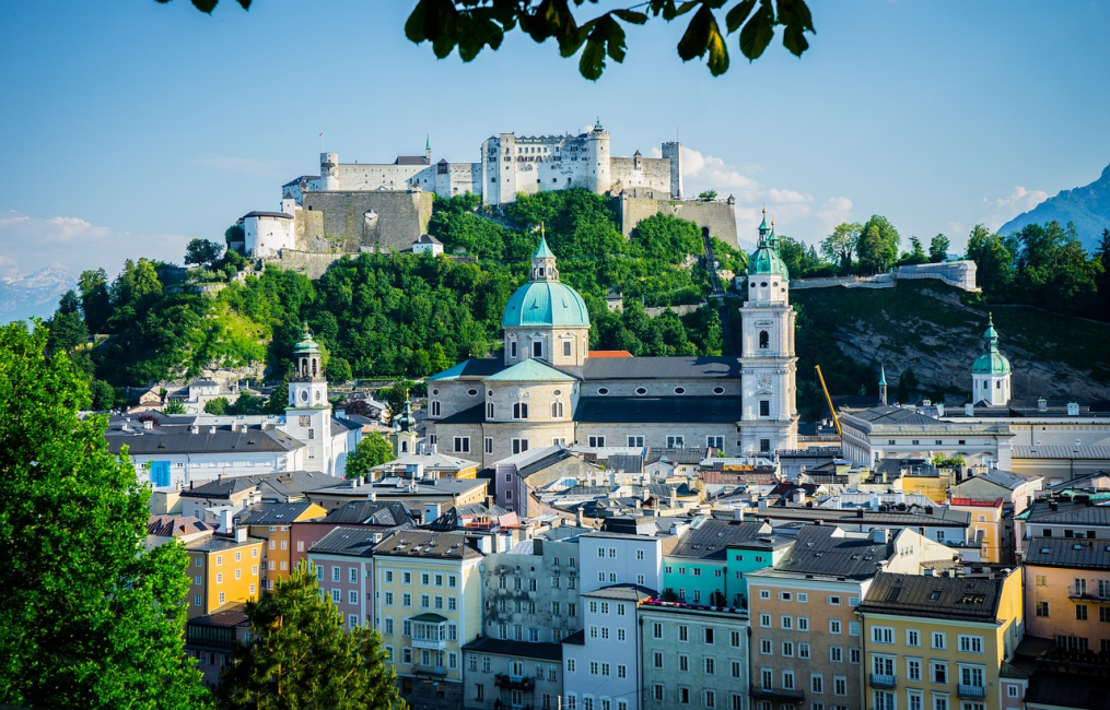 Yemyeşil ağaçlar ve berrak mavi gökyüzü altında, Hohensalzburg Kalesi'nin zirvede tüm heybetiyle yükseldiği, turkuaz kubbeli Salzburg Katedrali'nin ve altındaki çeşitli renklerdeki tarihi binaların sıralandığı canlı bir şehir manzarası.