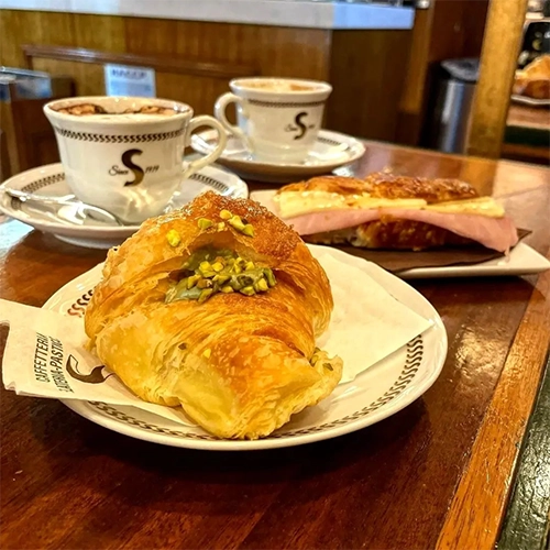 Sciascia Caffè'nin sıcak atmosferini yansıtan ahşap bir masada, ön planda Antep fıstığı dolgulu ve üzerinde fıstık parçacıkları olan çıtır bir kruvasan, arka planda ise üzerinde S 1919 logosu bulunan fincanlarda köpüklü iki kahve ve bir salamlı peynirli kruvasan iştah açıcı bir Roma kahvaltısını veya mola anını gözler önüne seriyor.