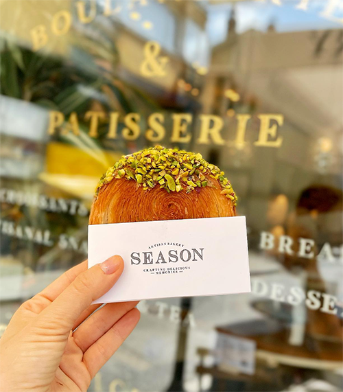 Bir el, beyaz Season Artisan Bakery logolu bir kılıf içinde, üzeri bolca doğranmış Antep fıstığı ile kaplanmış, kat kat, altın rengi, lezzetli bir kruvasan tutuyor. Arka planda camdan görünen bulanık vitrin yazıları, buranın bir pastane ve fırın olduğunu çağrıştırarak Bebek'in en iyi mekanlarından Season Artisan'ın özel fırın ürünlerini sergiliyor.