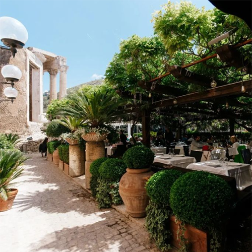 Bir Roma seyahat rehberi alternatiflerinden olan Tivoli'deki Ristorante Sibilla'da, beyaz örtülü masaların altında yemek yiyen misafirlerin bulunduğu yemyeşil pergolalı terasın yanı sıra, antik sütunları ve taş yapılarıyla büyüleyici tarihi kalıntıların gün ışığıyla aydınlandığı huzurlu bir dış mekân restoranı görülüyor.