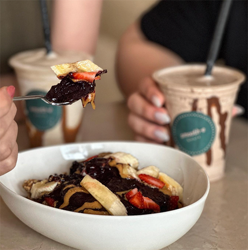 Koyu renkli, sağlıklı bir smoothie kasesinden muz, çilek ve fıstık ezmesiyle süslenmiş bir lokmanın kaşıkla alındığı bu yakın çekimde, arka plandaki Smooth-e & More logolu içecekler Bebek'in en iyi mekanları arasında yer alan bu mekanda sunulan ferahlatıcı ve besleyici lezzetleri sergiliyor.