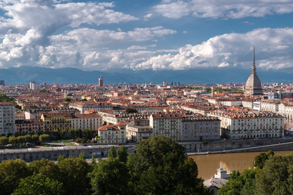 Mavi gökyüzü ve bulutların altında, uzakta belirginleşen dağ silsilesiyle çevrili Torino şehrinin geniş panoramik görüntüsünde, ön planda yemyeşil ağaçlar, kahverengi nehir kıyısı ve kiremit çatılı binalar arasında sivri kubbeli Mole Antonelliana dikkat çekiyor.