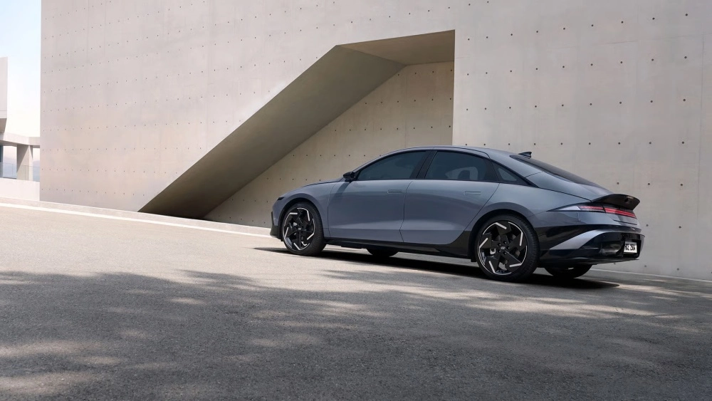 Koyu gri yeni Hyundai Ioniq 6 elektrikli otomobil, arka yan profilinden şık ve akıcı aerodinamik tasarımıyla, minimalist beton bir yapının geometrik detaylarla dolu modern cephesinin önünde asfalt yolda duruyor.