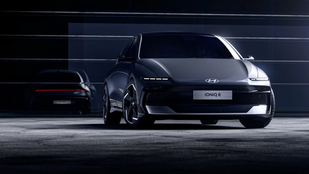 Ön planda belirgin LED gündüz farları ve IONIQ 6 logosuyla dikkat çeken, koyu gri renkteki yeni Hyundai IONIQ 6 elektrikli otomobilin modern ve aerodinamik dış tasarımı, loş ışıklı bir stüdyoda arkada belirsiz bir araç yansımasıyla sergileniyor.