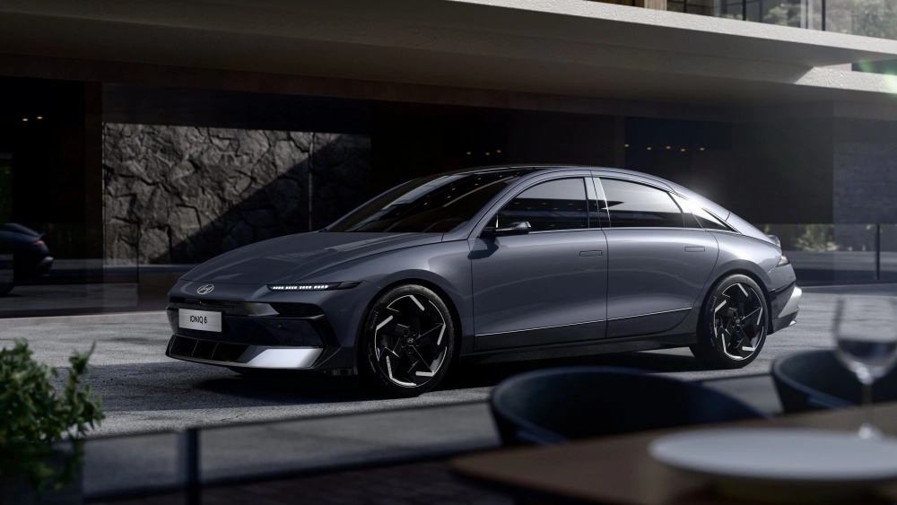 Yepyeni bir Hyundai Ioniq 6 elektrikli otomobil, koyu gri rengi ve keskin LED farlarıyla dikkat çeken modern ön tasarımıyla, lüks bir binanın önünde sol ön çaprazdan park edilmiş halde duruyor, dinamik ve geleceğe dönük yapısını sergiliyor.