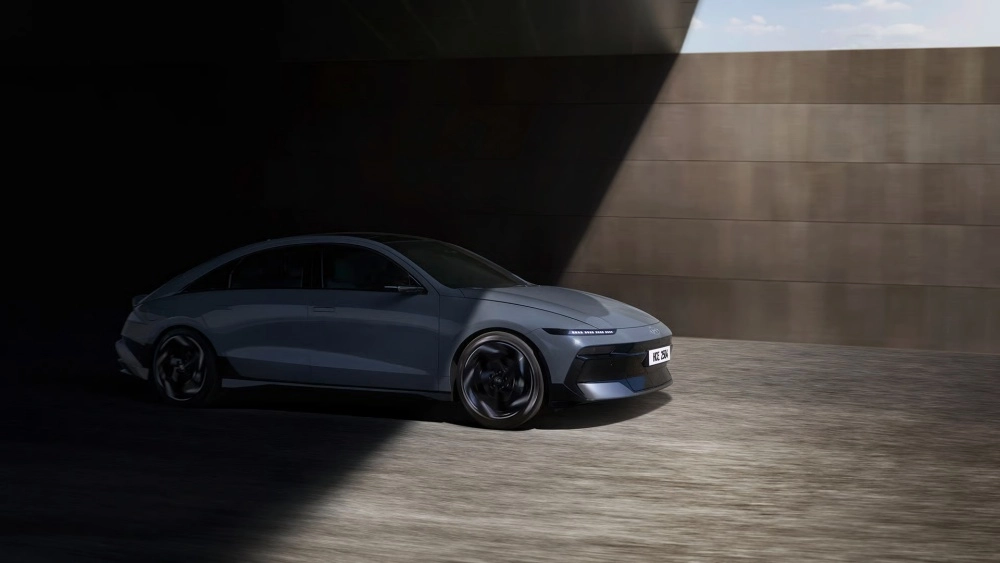 Gri renkli yeni Hyundai Ioniq 6 elektrikli otomobil, aerodinamik ve futuristik ön tasarım detaylarını ortaya koyan güçlü bir ışık hüzmesiyle aydınlatılmış dinamik bir sahnede, beton bir duvarın önünde durarak 680 km menzil kapasitesini hissettiriyor.