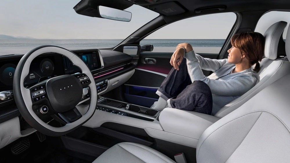 Yeni Hyundai Ioniq 6'nın konforlu iç mekan tasarımında, beyaz döşemeli yolcu koltuğuna uzanmış bir kadın deniz manzarasına bakarak rahatlarken, modern kokpit ve ortam aydınlatması aracın şık ve teknolojik ambiyansını vurguluyor.