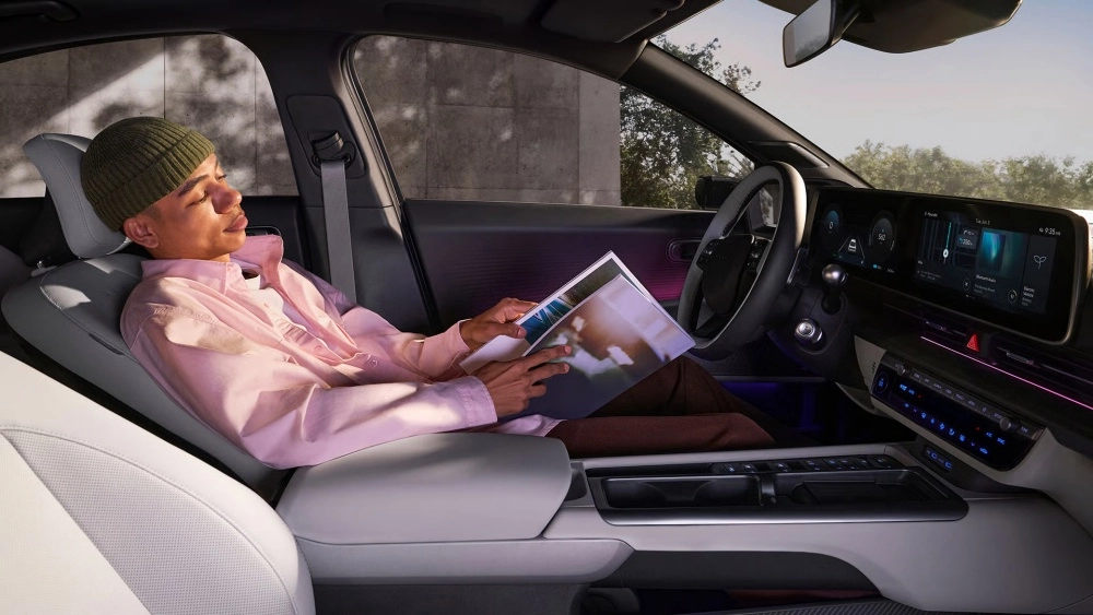 Hyundai Ioniq 6'nın ferah iç mekan tasarımında, yolcu koltuğunda elinde dergiyle rahatlamış bir kişi görülüyor; modern ön konsoldaki geniş dijital ekran ve şık ambiyans aydınlatması elektrikli otomobilin sunduğu konforu vurguluyor.