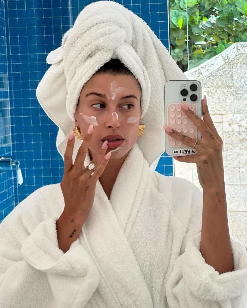 Beyaz havluya sarılı saçları ve yine beyaz bornozuyla aynadan selfie çeken bir kadın, yüzüne nemlendirici krem sürerken, pembe ojeli ve gösterişli yüzüklü eliyle cildine özen gösterdiği anı yansıtıyor; bu doğal ve ışıl ışıl görünüm, makyajsız makyajın ve sağlıklı cilt bakımının ön planda olduğu Vanilla Girl estetiğinin temiz ve ferah güzellik anlayışını mükemmel şekilde vurguluyor.