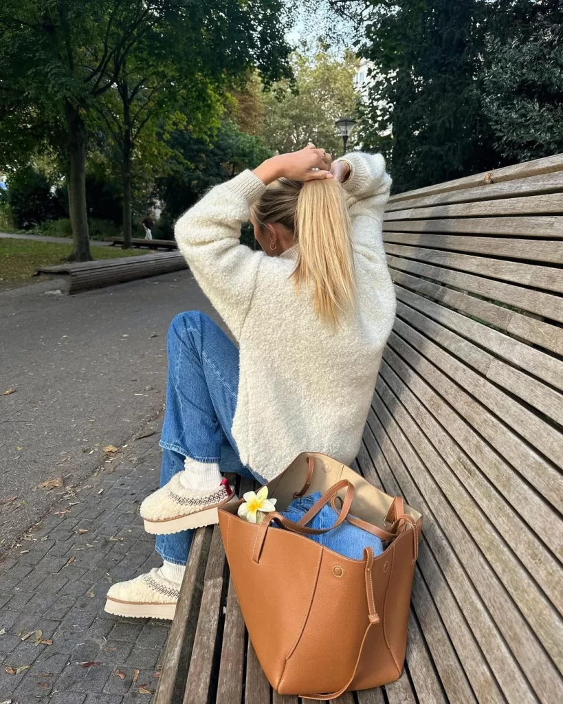Bir park bankında sırtı dönük oturan, vanilya kız estetiğini yansıtan sarı saçlı bir kadın, krem rengi kabarık kazağı, kot pantolonu, desenli beyaz çorapları ve platform tabanlı tüylü terlikleriyle saçını toplarken, yanında duran büyük kahverengi çantasında vanilya rengi bir çiçek göze çarpıyor ve bu sıcak görünüm soğuk sonbahar günlerine neşe katıyor.