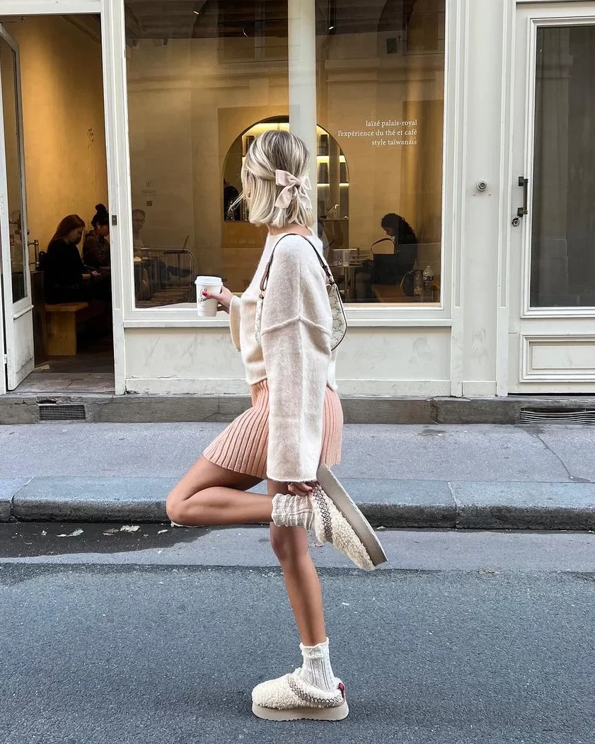 Sarışın, saçında krem fiyonk olan bir kadın, Vanilla Girl estetiği ruhunu yansıtan oversize krem örgü kazağı, fitilli açık pembe mini eteği, sıcacık örgü çorapları ve tüylü platform Ugg terlikleriyle sokakta kahve bardağıyla şık ve rahat bir şekilde yürüyerek soğuk sonbahar günlerine uygun, konforlu ve natürel bir şıklık sunuyor.