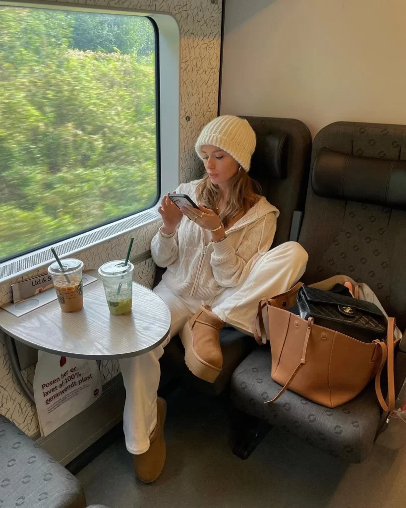 Tren koltuğunda oturmuş, telefonuna bakarken pencereden hızla geçen yemyeşil doğanın keyfini çıkaran, krem rengi örgü beresi, hırkası ve platform Ugg botlarıyla sıcak bir Vanilla Girl estetiği sergileyen genç bir kadın, yanında iki kahve ve çantalarıyla rahat bir yolculuk anını yansıtıyor.