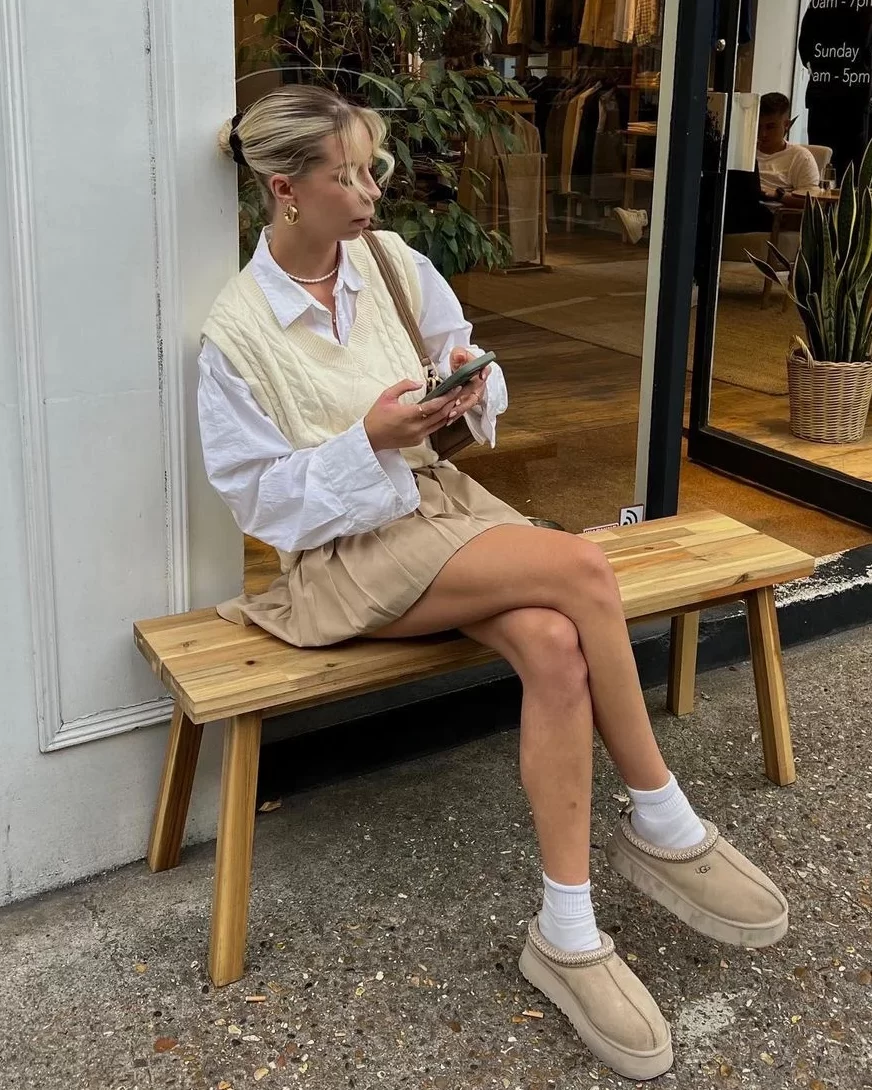 Bir bankta oturan, vanilya tonlarında örgü yelek, beyaz gömlek ve bej pileli mini etek giymiş bir kadın, Vanilla Girl estetiğinin sade ve konforlu kış stilini yansıtan bej UGG terlikleri ve beyaz çoraplarıyla telefonuna bakıyor.