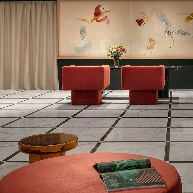 Alcron Prague Hotel'in lobisinde, geometrik desenli mermer zeminde, iki kırmızı koltuk, yuvarlak bir puf, ahşap sehpa ve duvarı süsleyen soyut tablolar, Prag seyahatinizin lüks konaklama seçeneklerinden birini sergiliyor.
