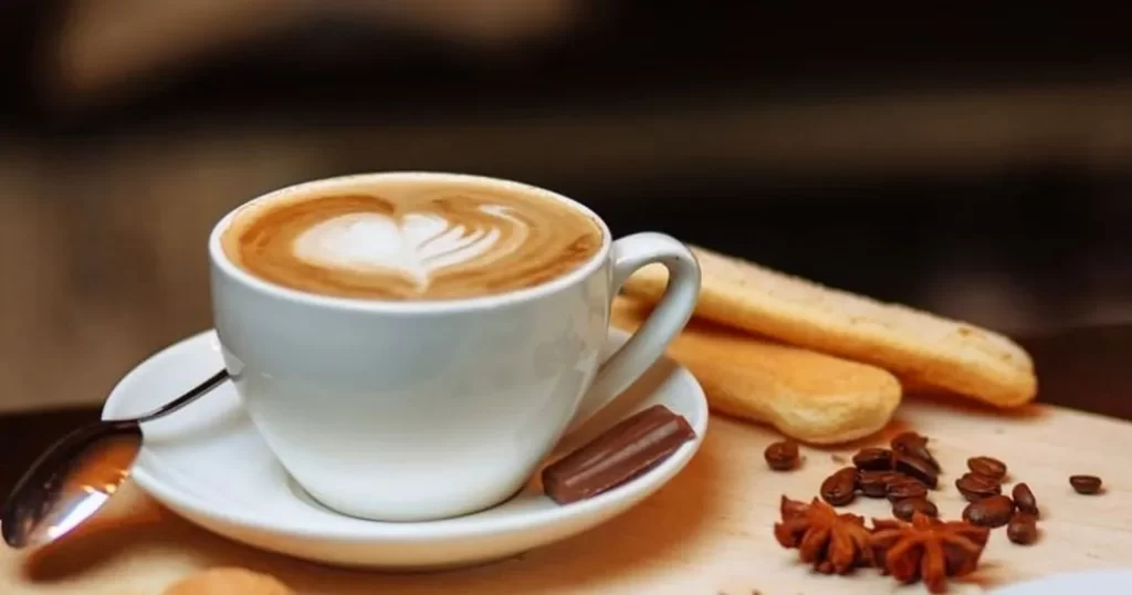 Şık beyaz fincanda kalp desenli latte köpüğüyle hazırlanmış bir kahve, ahşap bir zeminde tatlı kedi dili bisküvileri, kahve çekirdekleri ve yıldız anason baharatları eşliğinde, mistik güzelliğiyle ünlü Prag seyahatinin en keyifli kahve molasını sunuyor.