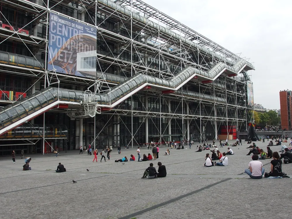 Paris'teki Centre Pompidou binasının dış cephesini saran çelik iskeletin ve cam tüp şeklindeki zikzak merdivenlerin önünde, Beaubourg Meydanı'nda toplanmış çeşitli insanlar, bazıları oturarak bazıları ise dolaşarak yoğun bir kalabalık oluşturuyor.