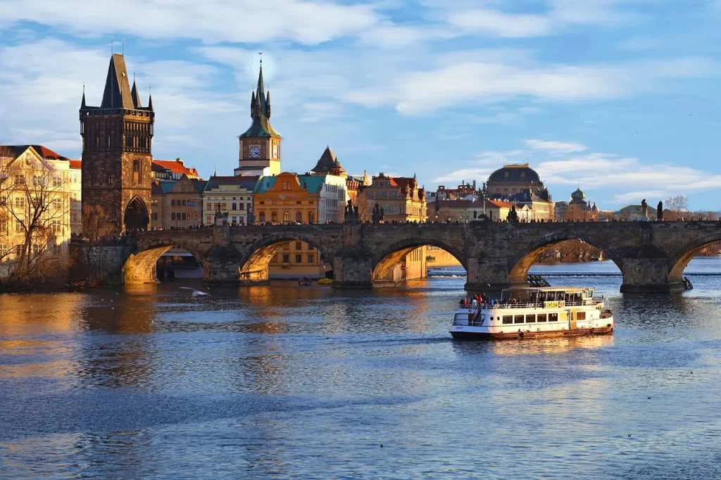 Vltava Nehri üzerinde, tarihi Charles Köprüsü'nün gotik kemerleri ve üzerindeki kalabalık siluetler, arkada Prag'ın mistik güzelliğini yansıtan eski kuleleri ve renkli yapılarıyla aydınlık bir havada, su üstünde süzülen bir tekne eşliğinde büyüleyici bir seyahat anı sunuyor.