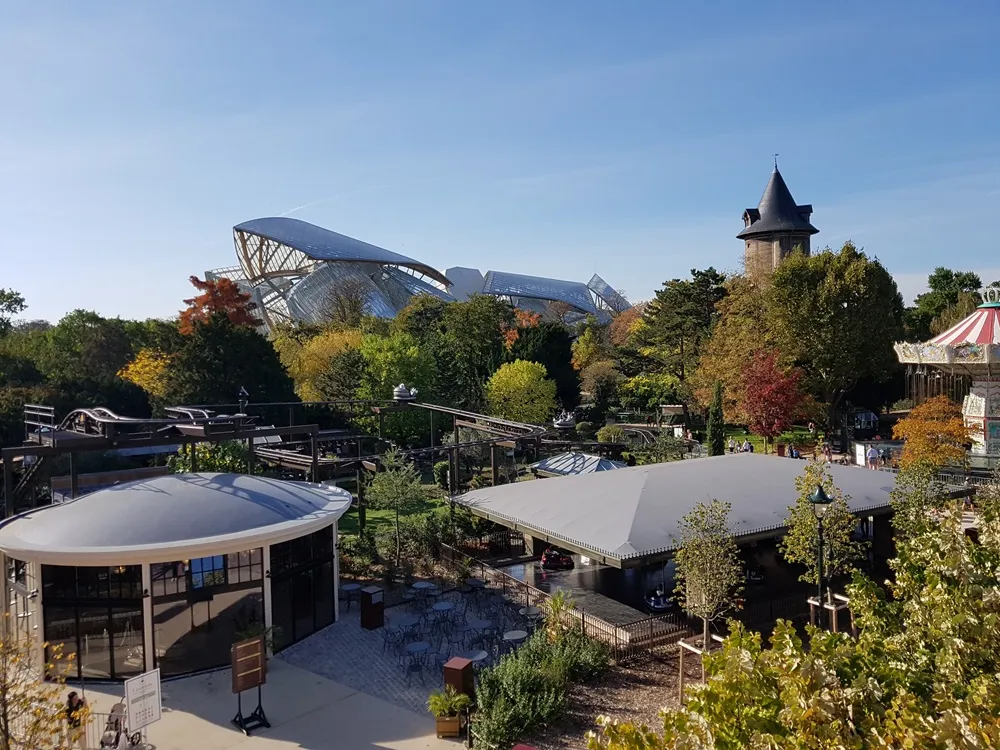 Canlı sonbahar renkleri sergileyen bir parkın ardında, kendine özgü modern tasarımıyla Fondation Louis Vuitton binası yükselirken, parkta sivri çatılı geleneksel bir kule, renkli bir atlıkarınca, eğlence parkı rayları ve ön planda açık havada oturma alanları olan çağdaş pavyonlar berrak gökyüzü altında uzanıyor.