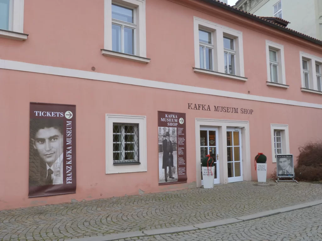 Açık şeftali rengi dış cephesi, beyaz çerçeveli pencereleri, ana giriş kapısının üzerindeki KAFKA MUSEUM SHOP yazısı ve Franz Kafka’nın portrelerini taşıyan bilgilendirme panolarıyla Mistik Güzelliğiyle Prag seyahat rehberi kapsamında ziyaret edilecek önemli noktalardan Franz Kafka Müzesi’nin ön cephesi, taş döşeli bir yoldan görülüyor.