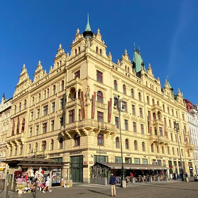 Mistik güzelliğiyle Prag seyahati deneyimini zenginleştiren, Art-nouveau tarzı zengin detaylı şık mimarisiyle öne çıkan, Kings Court Otel tabelalarının asılı olduğu görkemli yapının kafe ve restoran teraslarında günün keyfini çıkaran insanlarla dolu bir cadde sahnesi.